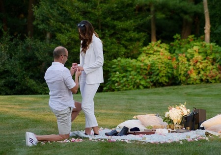James Murray down on one knee putting a ringer on his girlfriend's finger during an outdoor picnic.